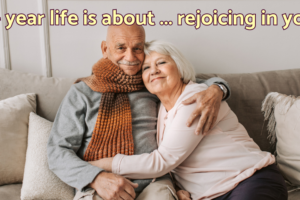 An elderly white male-female couple sitting on a couch and hugging. Text over the image reads "A 100+ year life is about ... rejoicing in your age."